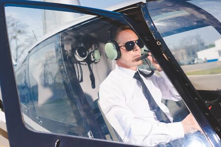Handsome mature Pilot in sunglasses and headset sitting in helicopter cabinの写真素材