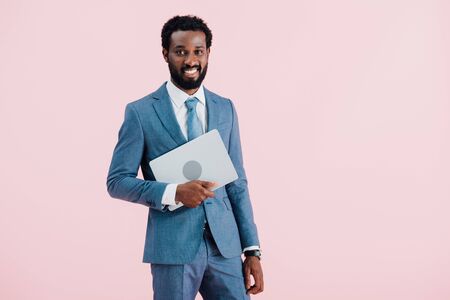 Cheerful African American businessman holding laptop isolated on pink backgroundの写真素材