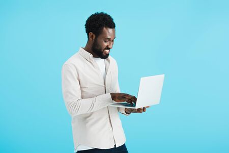 Smiling African American man in white shirt using laptop isolated on blue background