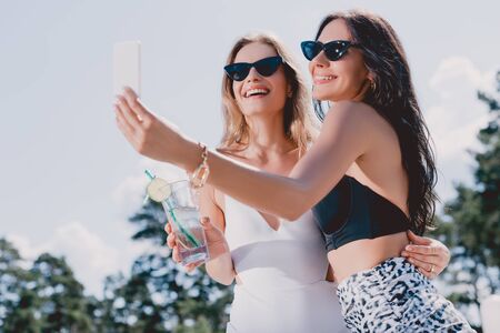 Low angle view of happy brunette and blonde women in swimsuits taking selfieの写真素材