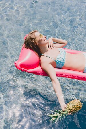 overhead view of wet happy blonde woman swimming on pink pool float with pineapple in swimming pool