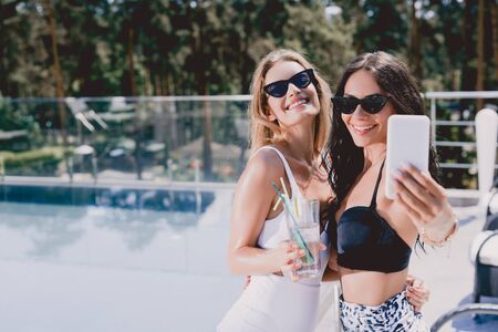 Sexy and smiling brunette and blonde women in swimsuits taking selfie near swimming pool