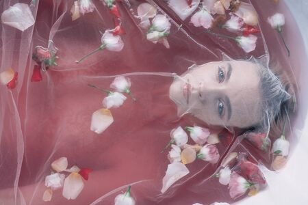 top view of beautiful woman covered with white mesh cloth in pink water with rose petals