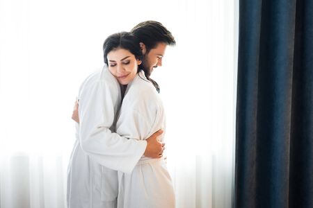 Cheerful bearded man hugging attractive woman in hotel roomの写真素材