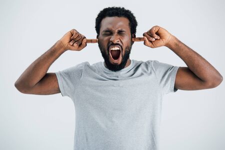 Emotional African American man in grey t-shirt shouting and closing ears isolated on grey backgroundの写真素材