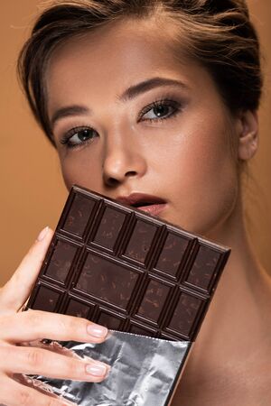 Beautiful young woman holding chocolate bar in silver foil near face isolated on beige background