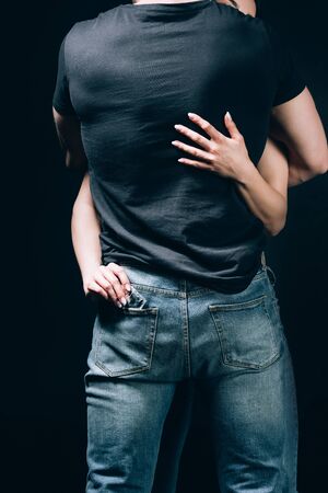 Back view of woman hugging man in jeans and taking out condom from pocket isolated on black backgroundの写真素材