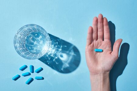 Partial view of woman holding blue pill on palm near glass with water on blue backgroundの写真素材