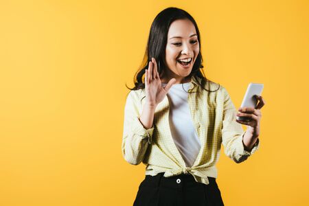 Happy Asian girl waving and making video call on smartphone isolated on yellow backgroundの写真素材