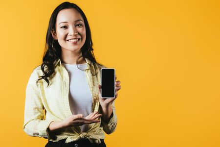 Attractive Asian girl presenting smartphone with blank screen isolated on yellow background