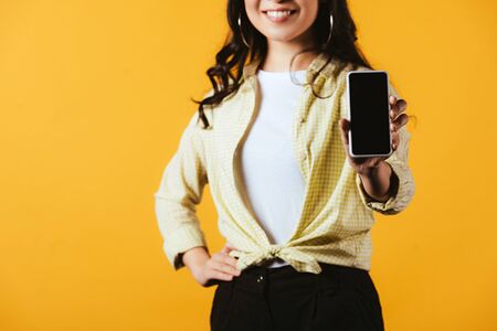 Cropped view of smiling girl showing smartphone with blank screen, isolated on yellow backgroundの写真素材