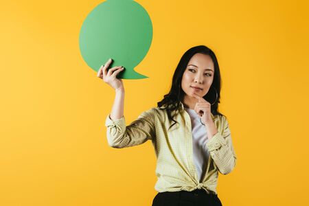 Thoughtful Asian woman holding green speech bubble, isolated on yellow backgroundの写真素材