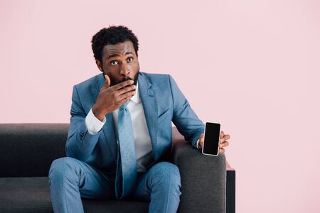 Shocked African American businessman sitting in armchair and showing smartphone with blank screen, isolated on pink backgroundの写真素材