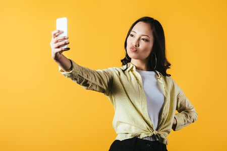 Attractive Asian girl taking selfie on smartphone isolated on yellow backgroundの写真素材