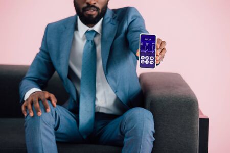 Cropped view of African American businessman sitting on armchair and showing smartphone with health app, isolated on pink backgroundの写真素材