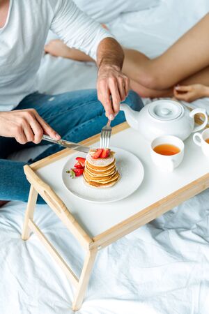cropped view of man eating pancakes with strawberries and drinking tea in bedの写真素材