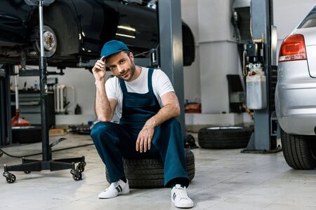 Happy bearded man touching cap and sitting on car tireの写真素材