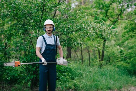 Cheerful gardener in overalls, helmet and hearing protectors holding telescopic pole saw and smiling...