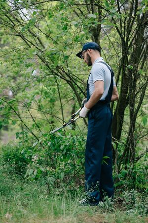 Gardener in overalls and cap cutting bushes with trimmer in garden