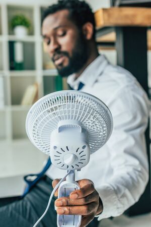 Selective focus of African american businessman sitting on floor and holding electric fanの写真素材