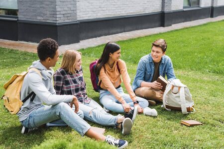 Smiling teenagers sitting on grass, talking, holding book outsideの写真素材