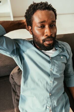 Unhappy African american man in sweaty shirt sitting on couch and suffering from summer heatの写真素材