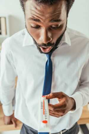 Shocked African american businessman looking at thermometer showing high temperature in officeの写真素材