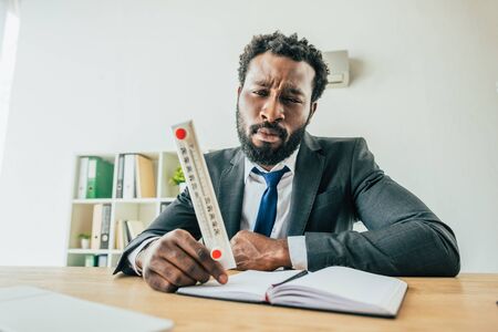 Unhappy African american businessman holding thermometer showing high temperature while sitting at workplace in officeの写真素材