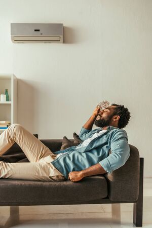 African american man lying on sofa with closed eyes and holding bottle with water near headの写真素材