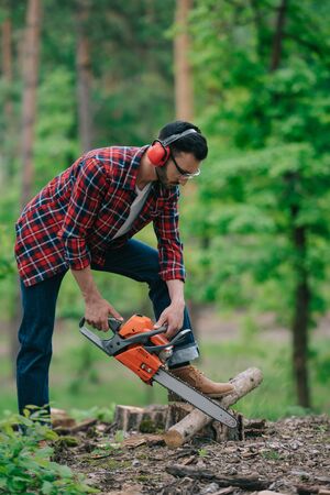 Lumberer in noise-canceling headphones cutting wood with chainsaw in forestの写真素材
