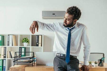 Dissatisfied African american businessman looking at sweaty shirt while suffering from summer heatの写真素材