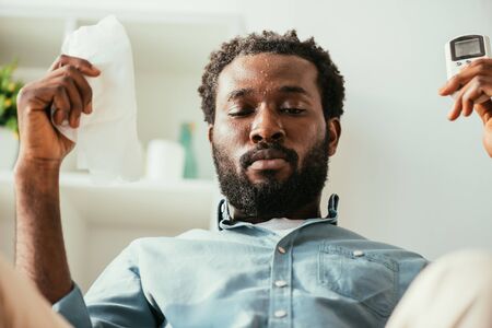 African american man with sweaty face holding napkin and remote controller while suffering from heat at homeの写真素材