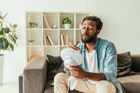 African american man holding blowing electric fan while suffering from heat at homeの写真素材