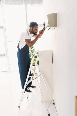Back view of African american repairman standing on ladder and fixing air conditionerの写真素材