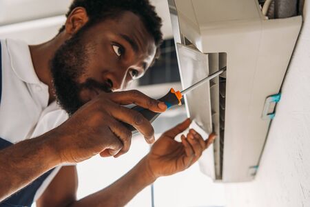 Serious African american repairman fixing air conditioner with screwdriverの写真素材