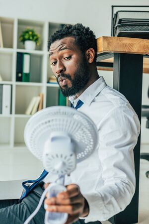Handsome African american businessman holding blowing electric fan while sitting on floor in officeの写真素材