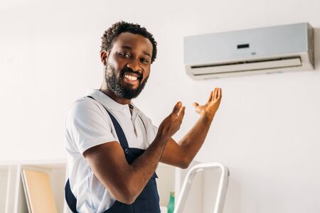 Cheerful African american repairman smiling at camera and pointing with hands at air conditionerの写真素材