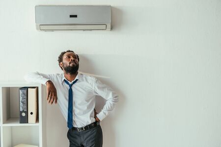 Young African american businessman standing under air conditioner and holding hand on hipの写真素材