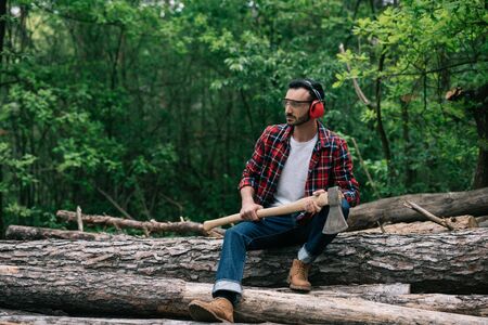 Pensive lumberman with ax sitting on logs in forest and looking awayの写真素材