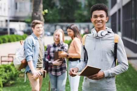 Smiling African american teenager in hoodie with headphones holding book and looking at cameraの写真素材