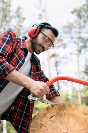 Cheerful lumberman in protective glasses and earmuffs cutting tree trunk with handsaw in forestの写真素材