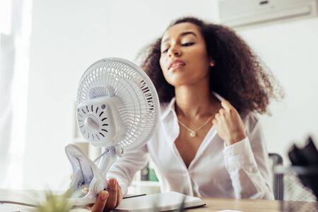 Selective focus of attractive African american businesswoman holding electric fan while sitting at workplaceの写真素材