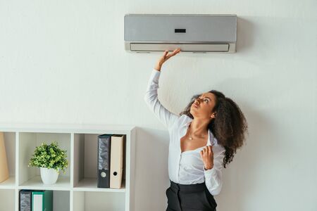 Pretty African american businesswoman touching air conditioner while suffering from heat in officeの写真素材