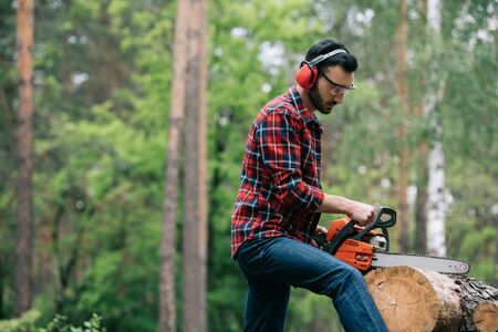 Attentive lumberman in hearing protectors cutting trunk with chainsaw in forestの写真素材