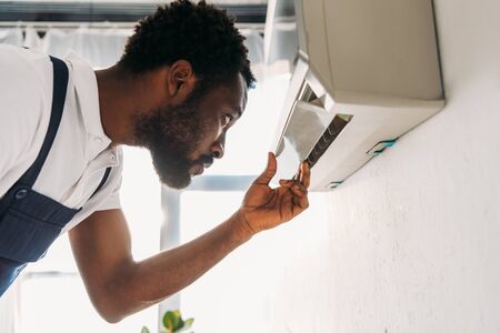 Concentrated African american repairman looking at broken air conditionerの写真素材