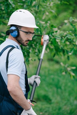 Selective focus of thoughtful gardener in helmet and hearing protectors trimming trees with telescopic pole saw in gardenの写真素材