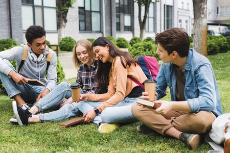 Smiling teenagers sitting on grass, talking, holding books and paper cupsの写真素材