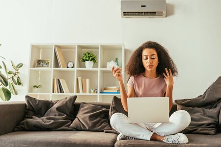 Serious African american woman using laptop and holding remote controller while sitting on sofa under air conditionerの写真素材