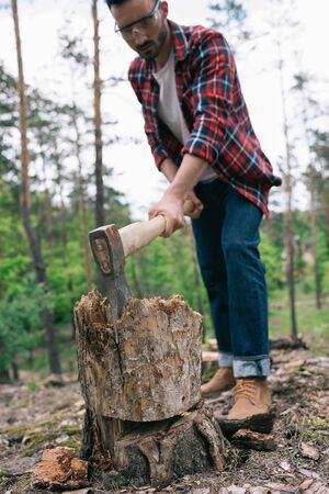 Selective focus of lumberman in plaid shirt and denim jeans cutting wood with ax in forestの写真素材
