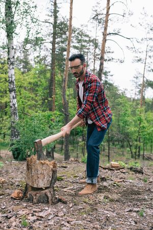 Handsome lumberjack in plaid shirt and denim jeans cutting wood with ax in forestの写真素材